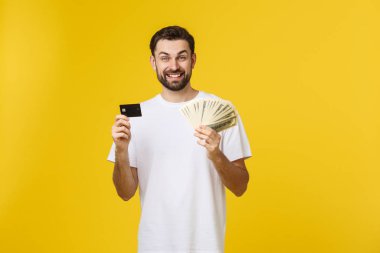 Genç, yakışıklı, mutlu, gülümseyen adam elinde bankacılık kartı ve para ile sarı arka planda izole bir şekilde..