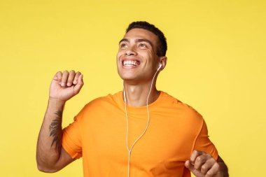 Neşeli, kaygısız bir adam gündüz yürüyüşünde mükemmel güneşli bir günün tadını çıkarıyor, bir elini kaldırıyor, neşeli bir şekilde dans ediyor, gülümsüyor, kulaklıkla mükemmel sesin tadını çıkarıyor, müzik kulaklıkları dinliyor, sarı arka plan.