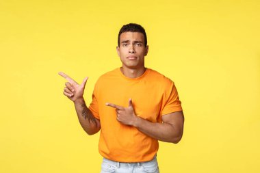 Sad gloomy cute masculine guy feeling offended or lonely, pointing left, sulking as looking camera disappointed, jealous, standing uneasy and distressed over yellow background, unhappy emotions