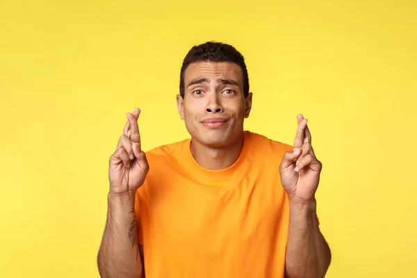 Hopeful worried young handsome man praying for wish come true, smirk silly and nervous, frowning as cant wait hear important results, want apply job, cross fingers good luck, yellow background — Stock Photo, Image