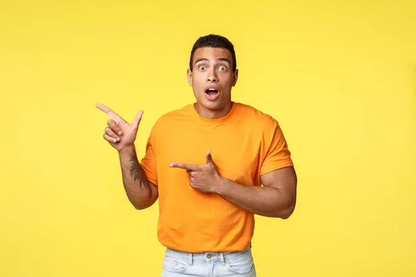 Shocked speechless young excited man seeing something fantastic or incredible, pointing upper left corner to attract attention, stare camera amazed, promote banner or product, yellow background — Stock Photo, Image