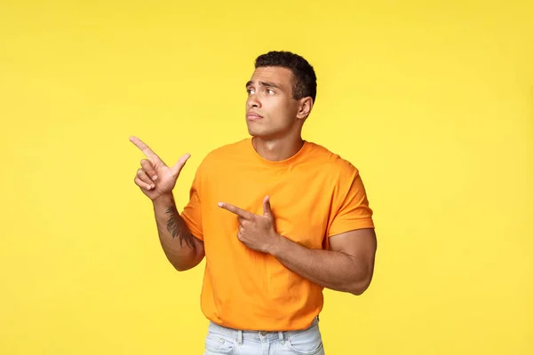 Ciúmes e chateado sombrio homem masculino bonito, use camiseta laranja, de pé infeliz e triste sobre fundo amarelo, apontando e olhando para a esquerda com pesar e sentimentos desconfortáveis, fundo amarelo — Fotografia de Stock