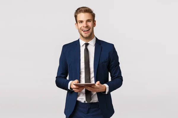 Alegre, elegante hombre de negocios barbudo en traje clásico y corbata, sosteniendo tableta digital riendo y sonriendo entusiasta, teniendo plan de negocios dispositivo escrito, de pie fondo blanco preparar discurso —  Fotos de Stock