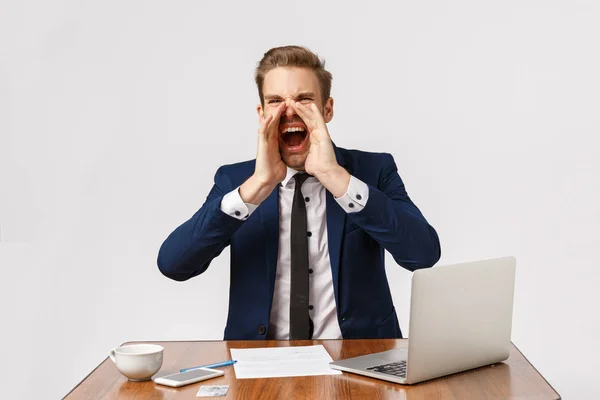 Expressive and emotional young blond man sitting in office, feeling pressured. Businessman with bad temper shouting out loud, calling someone, holding hands near mouth make voice louder — Stock Photo, Image