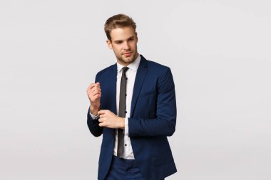 Classy good-looking blond bearded young male entrepreneur in suit, adjusting sleeves of jacket and looking away with pleased smile, relish good deal, knows how achieve success and money