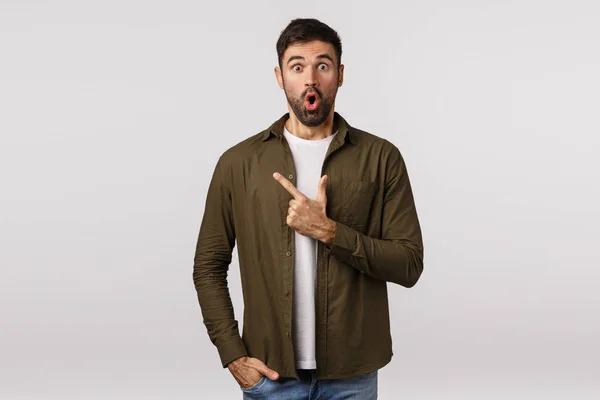 Surprised and amazed speechless young bearded man in coat, folding lips gasping impressed, pointing upper left corner astonished, heard interesting event taking place nearby, discuss news — Stock Photo, Image
