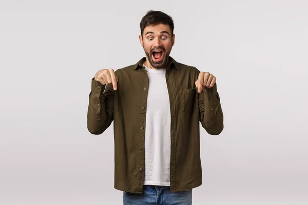 Animado, feliz e alegre bonito barbudo homem de casaco, apontando para baixo e olhando publicidade inferior com fascinado, expressão de alegria como ver promo Natal maravilhoso, fundo branco — Fotografia de Stock