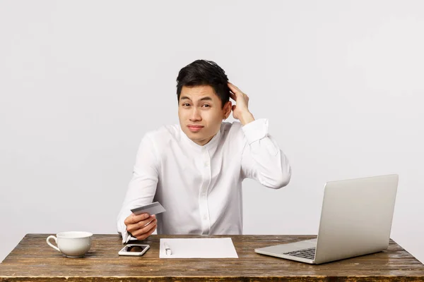 Confused, unsure young asian guy, office worker, sitting table near laptop, documents, scratching head hesitant, perplexed holding credit card, dont have cash suggest send money via bank