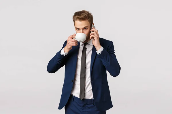 Нет времени на перерывы, сначала работа. Привлекательный молодой бизнесмен в классическом костюме, пьет кофе из чашки во время разговора по телефону, держит смартфон возле уха, звонит ассистенту взять одежду из прачечной — стоковое фото