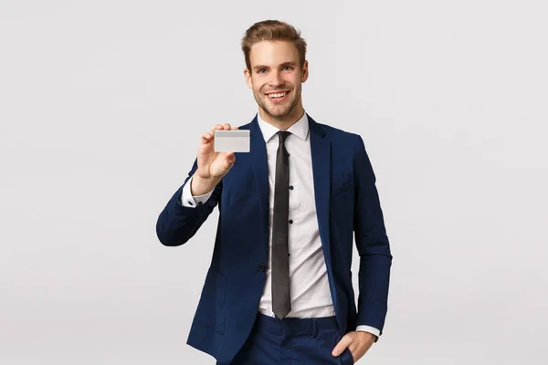 Gutaussehender, glücklicher erfolgreicher Geschäftsmann im klassischen Anzug, der mit Kreditkarte bezahlt, seine Bankkarte zeigt und zufrieden lächelt, betritt die Lounge-Zone des Flughafens, weißer Hintergrund stehend — Stockfoto