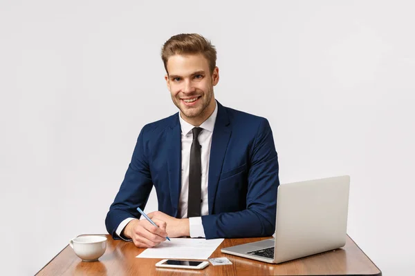 Zeit, Geld zu verdienen. gutaussehender Büroleiter sitzt an seinem Schreibtisch, schreibt Bericht und lächelt den Kunden an, hält Stift, bereitet Unterlagen vor, benutzt Laptop, trinkt Kaffee aus Tasse — Stockfoto