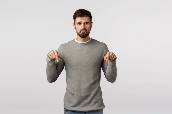 Gloomy and depressed, upset sulky boyfriend with beard in grey sweater, pointing down, pulling smile down distressed and jealous, feel regret or disappointment, standing white background — Stock Photo, Image