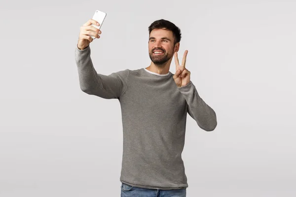 Optimistický a bezstarostný, šťastný atraktivní pejsek s vousy v šedém svetru, držící smartphone vzhůru jako selfie, aby mír znamení a úsměv, volání přítel video-hovor on-line, bílé pozadí — Stock fotografie