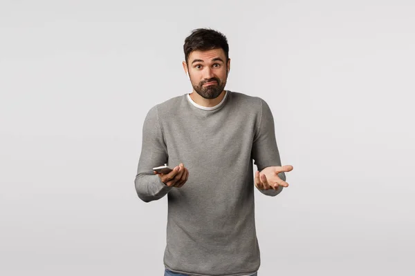 Unsure and clueless, puzzled handsome bearded male in grey sweater, feeling uncertain, smirk unaware, shrugging and raise hands sideways, holding smartphone, wear wireless earphones, white background — Stock Photo, Image
