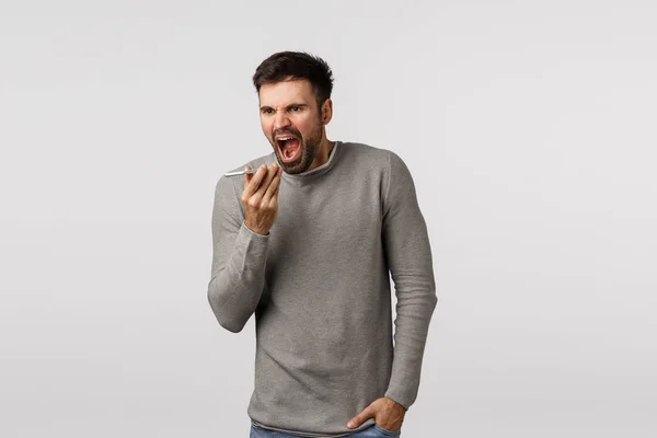 Angry, pissed boyfriend with beard lost temper, feeling pissed and outrated, complaining, arguing as record phone voice message using application, shouting at smartphone microphone, white background — Stock Photo, Image