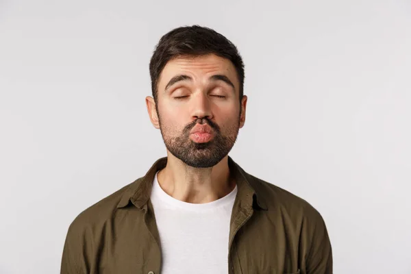 Chico barbudo guapo divertido y lindo en abrigo, ojos cerrados y labios plegables como esperando un beso apasionado, enviando mwah, teniendo cita romántica en el día de San Valentín, de pie fondo blanco — Foto de Stock