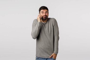 Kararsız ve kararsız sakallı, gri kazaklı, sırıtan, belirsiz, sorunlu cevap veren, el ele tutuşan, telefonla arkadaşını arayan, akıllı telefonu kulağının yanında tutan, beyaz arka planda duran sevimli eşcinsel adam.