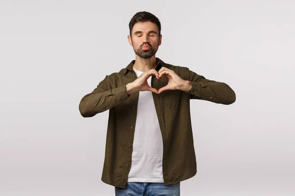 Bonito atraente e bonito jovem gay homem com barba em roupa moderna, fechar os olhos e puxar os lábios, bebendo para beijar, fazer gesto de coração, expressar carinho, parceiro de amor, pé fundo branco — Fotografia de Stock