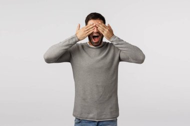 Neşeli ve kaygısız, gri kazaklı, mutlu, sakallı yakışıklı adam, avuçlarını kapalı, gülümseyerek merak ediyor, güzel bir sürpriz bekliyor, hediye görmek istiyor, peekaboo oynuyor, saklambaç oynuyor, beyaz arka plan.