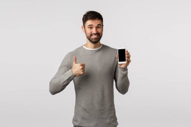 Gri kazaklı arkadaş canlısı, yakışıklı ve keyifli sakallı adam başvuru formu kullanmayı öneriyor ya da alışveriş sitesi, akıllı telefon tutmak, onaylı baş parmağını kaldırma, mutlu gülümseme gibi.
