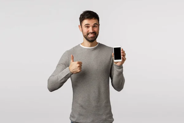 Přátelský pohledný a nadšený vousatý muž v šedém svetru doporučujeme použít aplikaci nebo jako nový messanger, nákupní stránky, držení smartphone, show thumb-up v souhlasu, jako, usměvavý potěšen — Stock fotografie