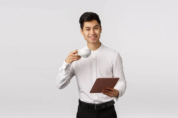 ¡Salud! Elegante hombre asiático guapo, empresario exitoso levantando taza de café y sonriendo complacido, sosteniendo tableta digital, aprobar documento, recibir respuesta positiva, fondo blanco — Foto de Stock