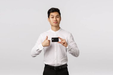 Yakışıklı Asyalı genç iş adamı banka tavsiye ediyor, kredi kartı tutuyor, başparmak kaldırıyor, sırıtıyor ve bir kaşını kaldırıyor gururlu, tatmin olmuş bir ifadeyle, beyaz arka planda duruyor.