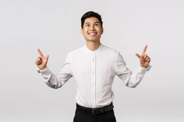 Vrolijke onder de indruk, dromerige knappe Aziatische man in shirt, zwarte broek, omhoog wijzend met tevreden, zorgeloze uitdrukking, zoals wat hij ziet, eens met banner, staande witte achtergrond — Stockfoto