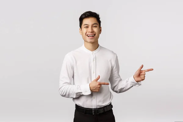 Carefree charismatické asijské mladé 25s muž ve formálním oblečení, bílá košile, černé kalhoty, pozvat check-out a vidět událost prapor, ukazující vpravo a usmívající se, smějící se pobavený, stojící bílé pozadí — Stock fotografie