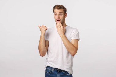 Şaşırmış ve şok olmuş genç sarışın yakışıklı adam, başparmağını sola doğru uzatıyor ve bakıyor, nefes nefese kalmış ağzı dedikodu gibi, şaşırtıcı bir olaya tanık oluyor, şok edici bir sahne, beyaz arka plan