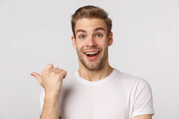 Surprised and amused excited, good-looking blond guy with bristle and blue eyes, smiling fascinated and amazed, pointing thumb left joyfully discuss good product, white background — Stock Photo, Image