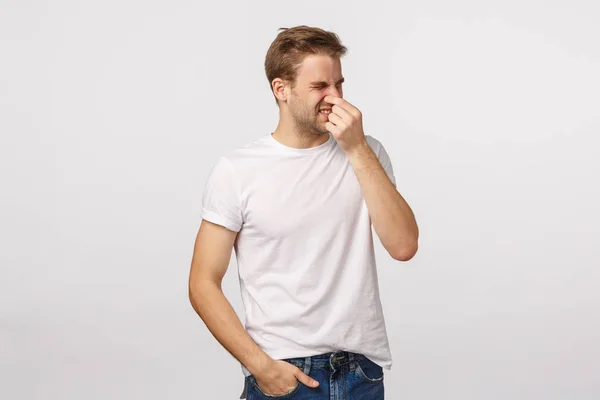 Disappointed and disgusted blond handsome caucasian man close nose with fingers, squinting and turn away from aversion and disgusted, smell something bad, product stinks, standing white background — Stock Photo, Image