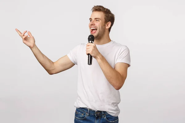 Happy charming blond bearded man in white t-shirt, reading lyrics from screen as attend karaoke with friends, holding microphone gesturing and smiling, singing favorite song, smiling white background — Stock Photo, Image