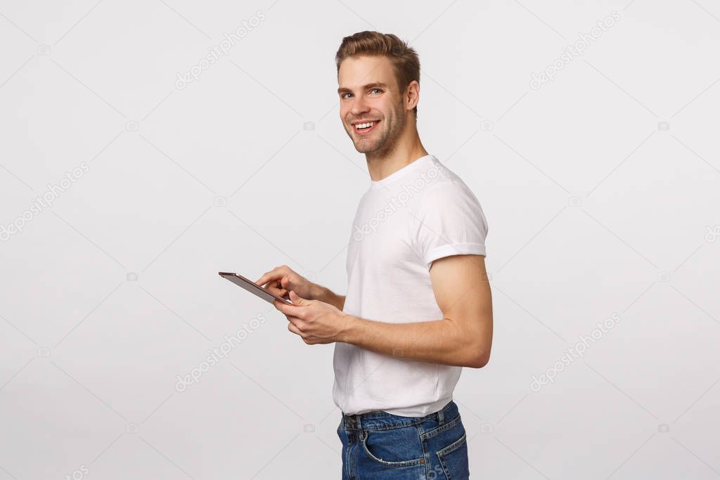 Attractive blond male office assistant, manager, using digital tablet to input or record information, using corporate application, standing profile, turn camera and smiling, white background