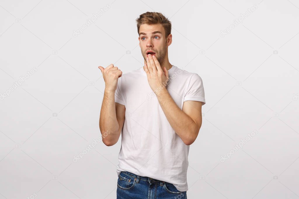 Surprised and shocked young blond handsome guy, ponting thumb left and staring, gasping cover mouth as gossiping, witness something astounding, shocking scene, standing white background