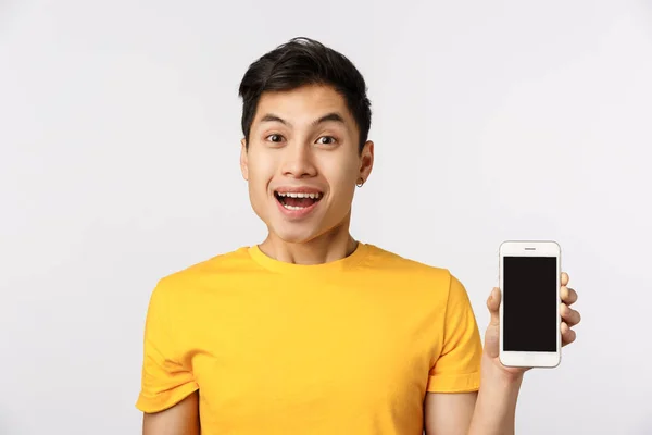 Deve ter no teu telemóvel. Alegre, animado jovem asiático homem em camiseta amarela, mostrando smartphone e sorrindo, adoro novos filtros no aplicativo, recomendo juntar-se à empresa, use recurso on-line, fundo branco — Fotografia de Stock