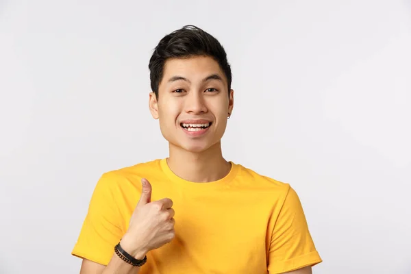 스튜디오를 클로즈업하면서 즐겁고 매력적 인 노란 티셔츠를 입은 아시아 남성을 찍었습니다. 엄지손가락을 들고 웃으면서 긍정적 인 피드백을 주고 회사 서비스를 추천하고, 선택을 승인하고, 백인 배경을 만들었습니다. — 스톡 사진