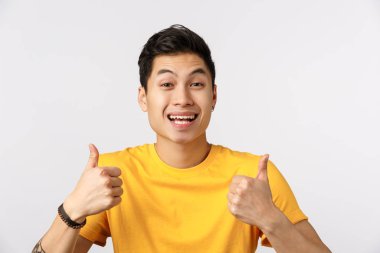 Yakın çekim, sarı tişörtlü çekici Asyalı adam devam etmenizi, başparmaklarınızı göstermenizi ve geniş bir gülümseme göstermenizi öneriyor. Fikri onaylamak, kavram gibi, arkadaşınızla iyi bir seçim yaptınız, beyaz arka plan.