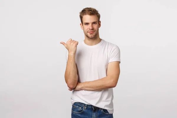 Hombre macho encantador asertivo con corte de pelo rubio, cerda, camiseta blanca, apuntando a la izquierda con el pulgar y sonriendo complacido, saber exactamente lo que necesita, recomendar lugar, empresa o producto, publicidad —  Fotos de Stock