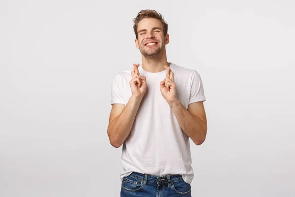 Pretty please god make my dream come true. Cute lucky and hopeful charming blond guy making wish, cross fingers good luck, smiling and raise head up sky, relish good news, white background — ストック写真