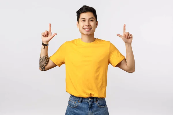 Veselý, přátelský a dobře vypadající asijský mladý student ve žlutém tričku, s tetováním, radostně se usmívající, ukazující nahoru, doporučující produkt, radící se tam, kde najdete vše, co potřebujete — Stock fotografie