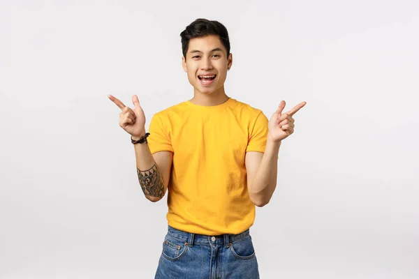 Bezczelny, przystojny azjatycki wytatuowany facet w żółtej koszulce, skierowany bokiem, pokazujący lewy i prawy wariant, dwa produkty, uśmiechnięty entuzjasta, stojące białe tło, sugerują możliwości — Zdjęcie stockowe