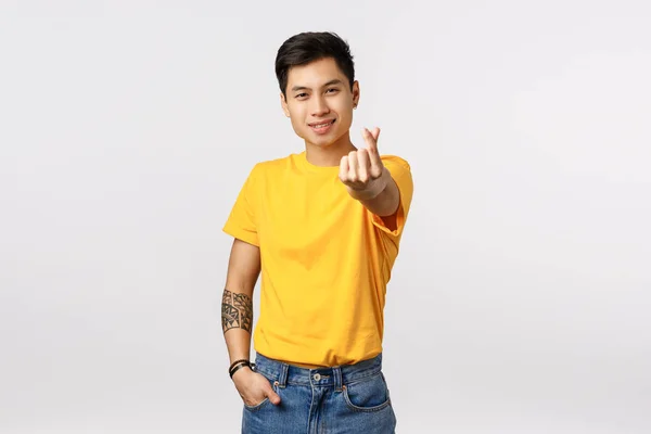 Bonito e elegante hipster asiático cara em amarelo t-shirt, com tatuagens, estender o braço e mostrando coreano coração amor sinal, sorrindo bobo, expressar carinho, adoro namorada, pé fundo branco — Fotografia de Stock