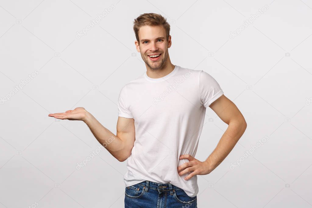 Cute smiling, happy young blond bearded man in white t-shirt introduce product, holding something on blank copy space, introduce chart or concept, grinning delighted, white background
