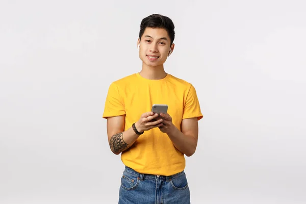 Knappe Aziatische man in geel t-shirt, jeans, draadloze oortjes dragen, smartphone vasthouden, muziek luisteren, afspeellijst maken in applicatie, karaoke zingen met behulp van telefoon app, witte achtergrond — Stockfoto