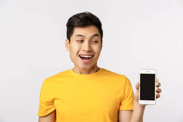 Guy realmente adorar novo aplicativo de filtro. Atraente asiático homem em amarelo t-shirt, mostrando smartphone display, olhando tela móvel e sorrindo entretido, fascinado com o quão incrível serviço funciona — Fotografia de Stock