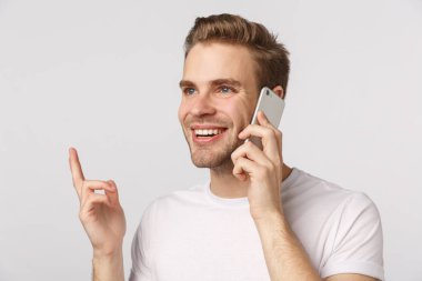 Yakın plan sarışın, mavi gözlü, beyaz tişörtlü, kulağının yanında akıllı telefon tutan, el kol hareketi yapan, gülümseyen, memnun olan, sipariş veren, online alışverişi onaylayan, arkadaşıyla konuşan, beyaz geçmişi olan yakışıklı bir adam.