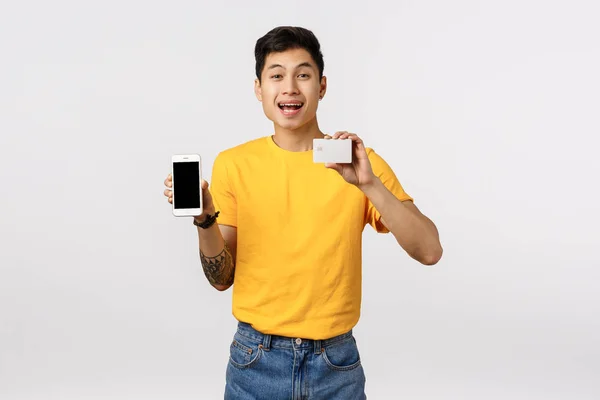 Щасливий усміхнений, веселий азіатський хлопець у жовтій футболці з татуюваннями, тримає смартфон і кредитну картку, рекомендує скористатися онлайн касовим апаратом, фінансовою системою, замовити продукт Інтернет, білий фон — стокове фото