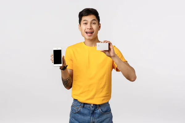 Sorrindo feliz, alegre asiático cara em camiseta amarela com tatuagens, segurando smartphone e cartão de crédito, recomendo o uso de cashback on-line, sistema financeiro, encomendar internet do produto, fundo branco — Fotografia de Stock