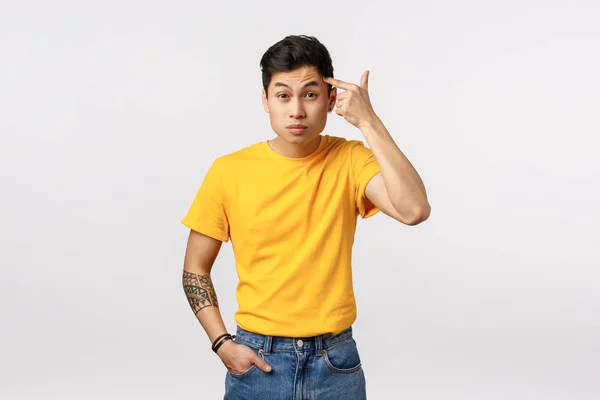 Maybe you should think before acting. Annoyed and bothered young asian guy in yellow t-shirt, with tattoos, pointing temple as scolding blaming someone being dumb or stupid, white background — Stock Photo, Image